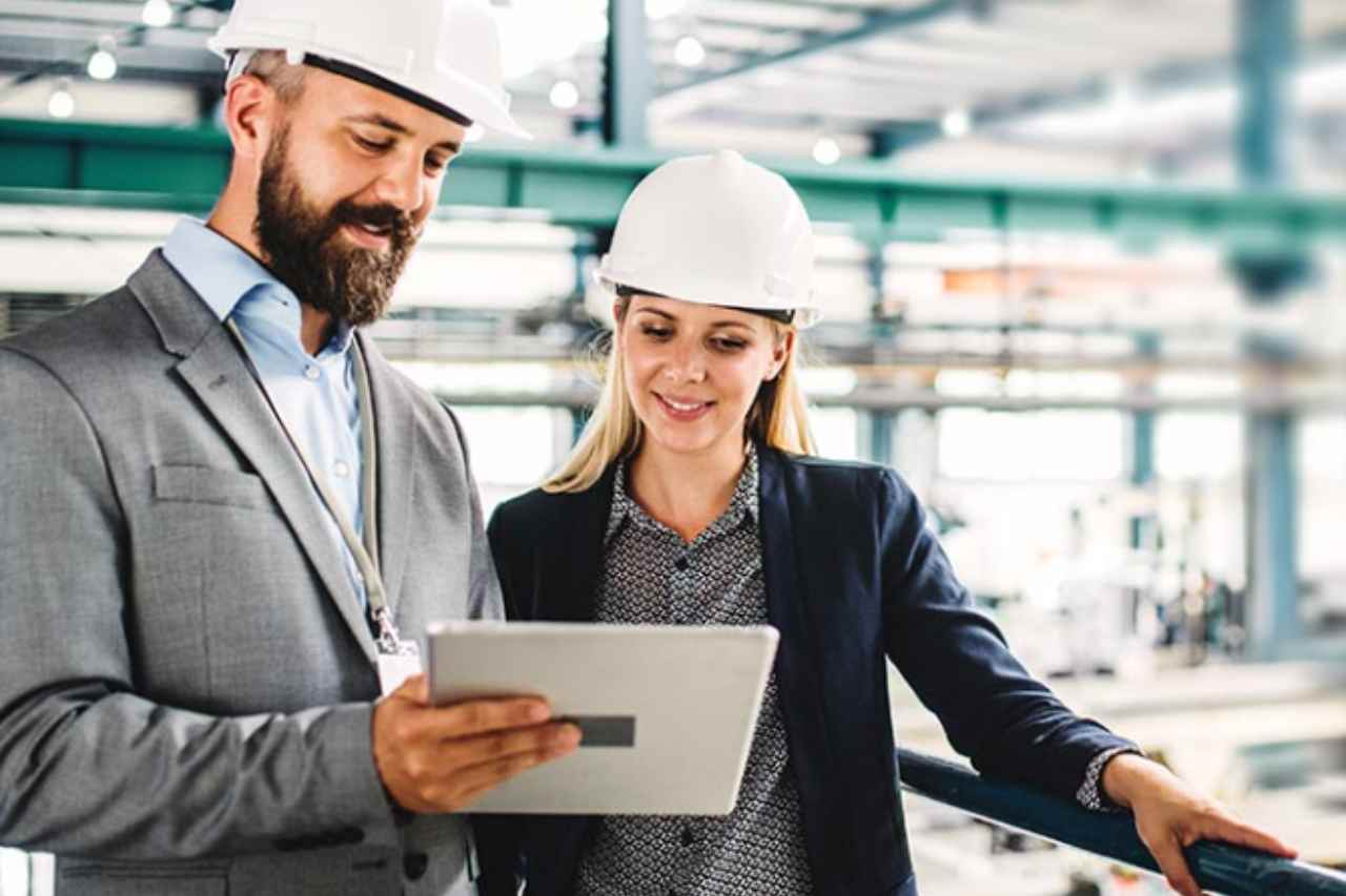 A man and woman in construction hats look at a digital tablet together in a warehouse.