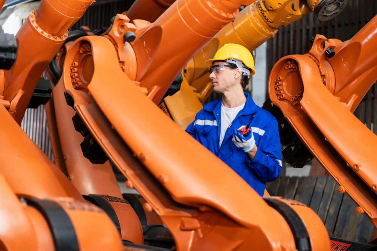 A manufacturing worker stands amongst large machinery in a factory wearing a hard hat.