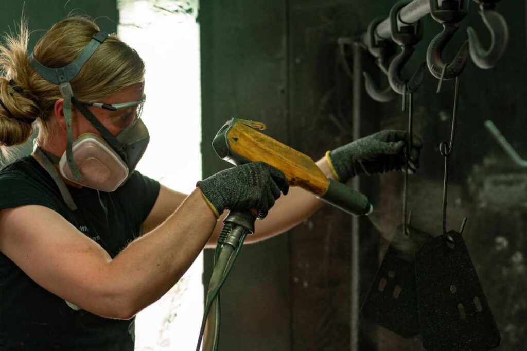 A woman in PPE uses a spray gun to apply powder coat paint.